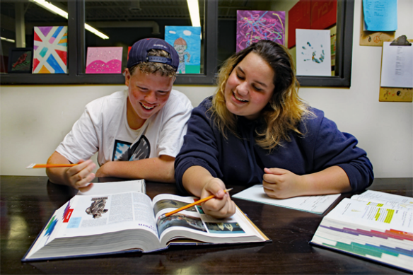 Two teenagers sitting together, looking down at a textbook and smiling, they are both holding pencils