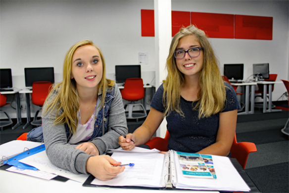 Two blonde teenage girls sit together in a computer lab with binders and papers as if they are studying, they are smiling at the camera