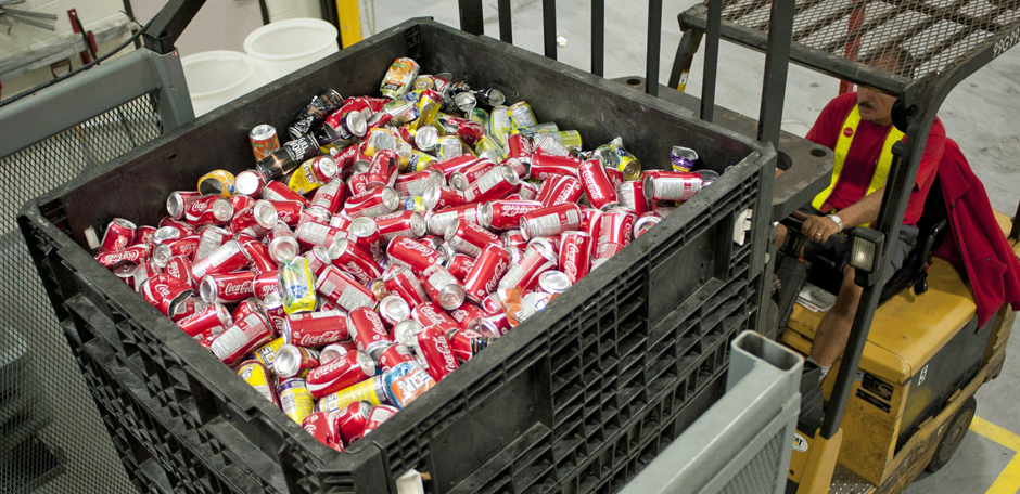 A man drives a forklift carrying up a large crate filled mostly with Coca-Cola cans, some other assorted cans are mixed in also